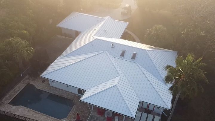 Aerial view of a white metal-roofed house with an attached pool in a wooded setting, captured during a hazy sunrise.