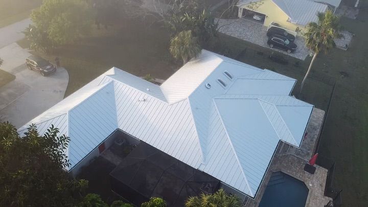 An aerial view of a light-colored metal roof on a house with a surrounding yard and pool.