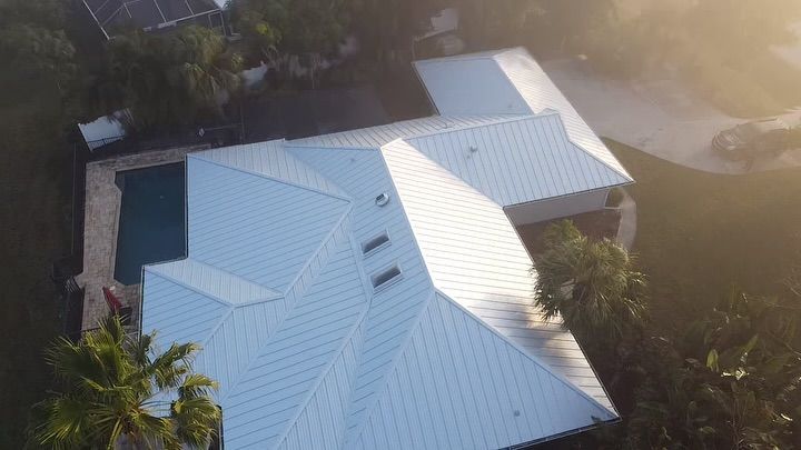 An aerial view of a residential house with a light-colored standing seam metal roof and a pool in the backyard.