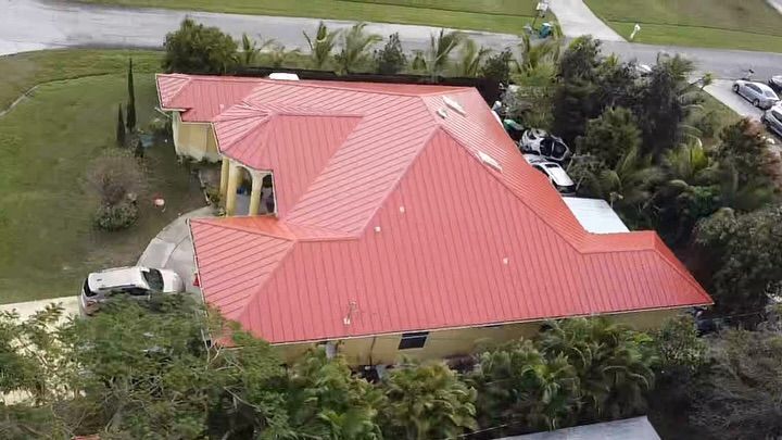 An aerial view of a yellow house with a bright red metal roof, surrounded by trees and vehicles in a residential area.