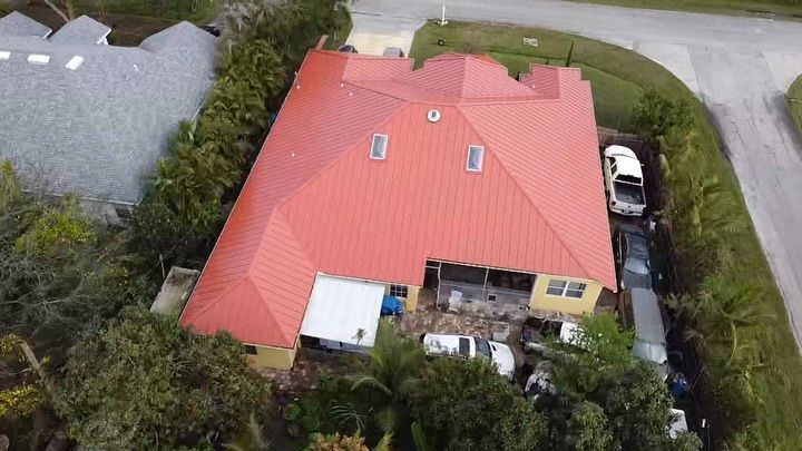 An aerial view of a suburban home featuring a bright red metal roof, surrounding trees, and vehicles parked in the driveway.