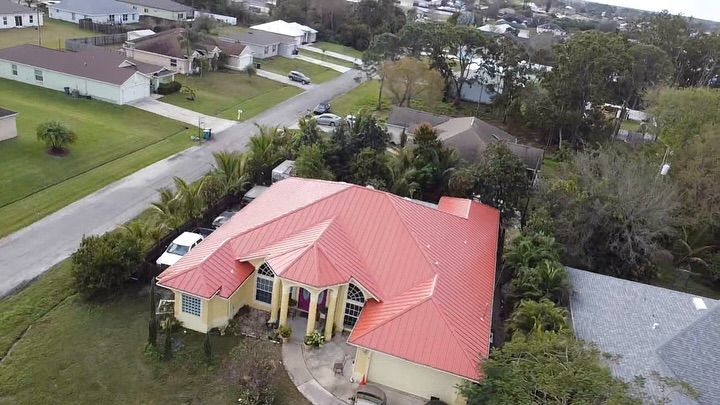 An aerial view of a suburban home with a bright red metal roof, surrounded by trees and neighboring houses on a street.