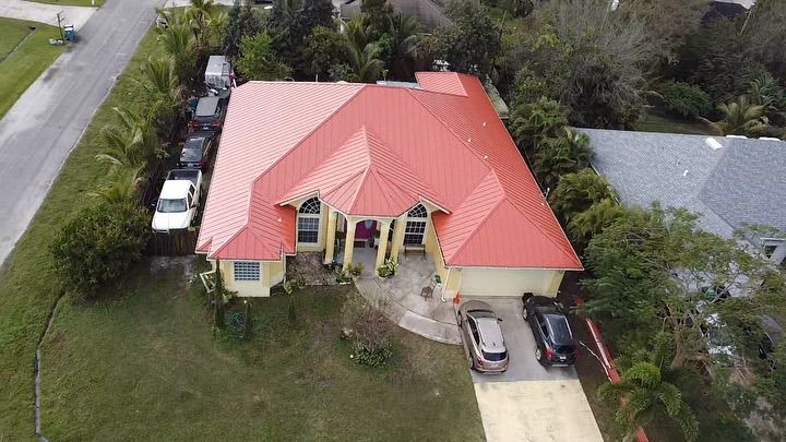 An aerial view of a yellow house with a bright red metal roof, surrounded by trees with cars parked in the driveway.