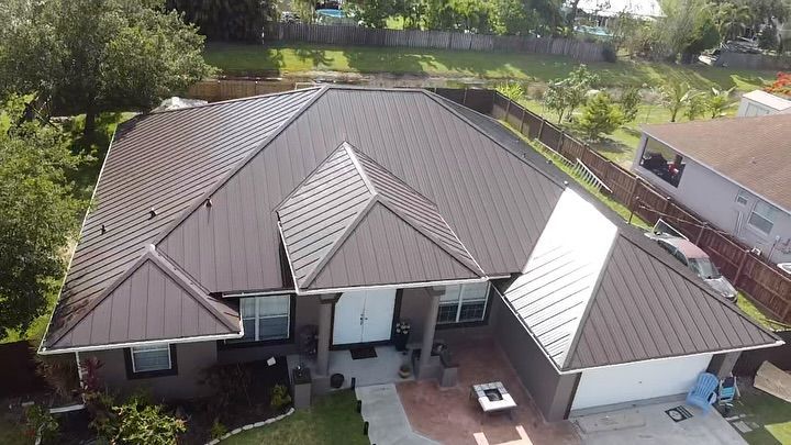A high-angle view of a residential house with a dark brown standing seam metal roof and a paved front entrance area.