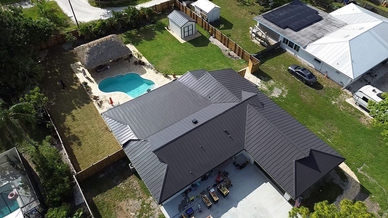 An aerial view of a residential house with a dark shingled roof, a backyard swimming pool, and a patio area.