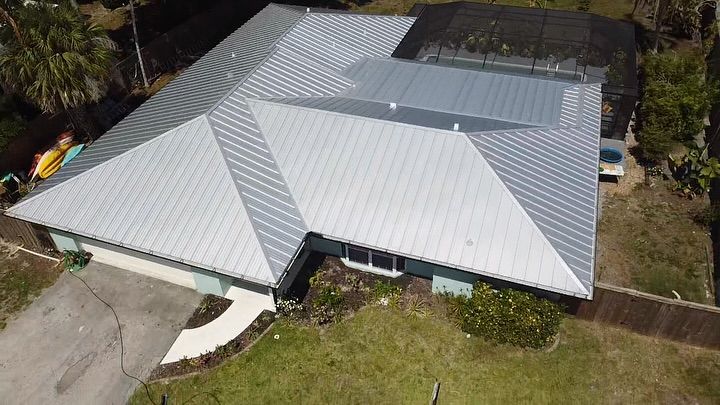An aerial view of a residential house featuring a light-colored metal roof, a driveway, and a screened-in pool enclosure.