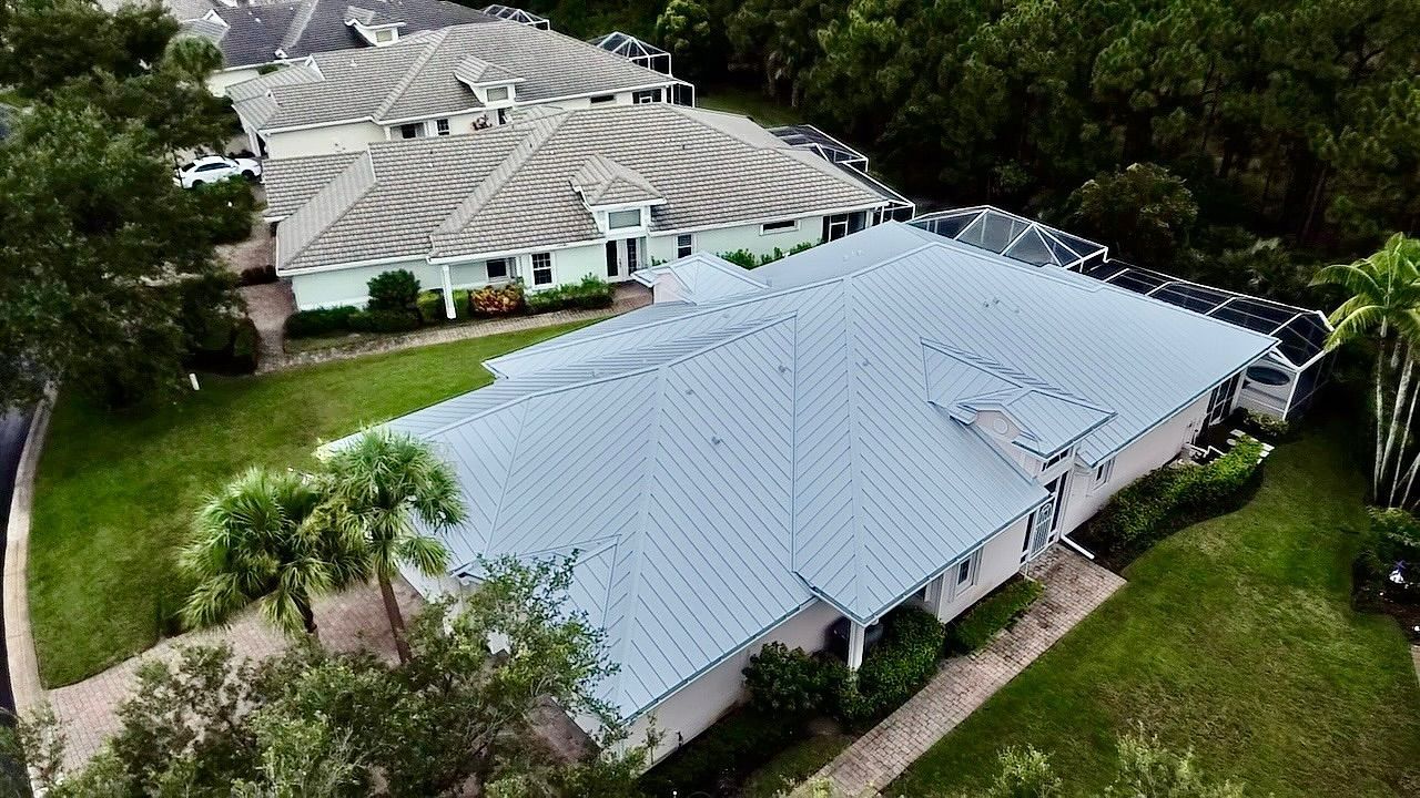 An aerial view of two residential houses with light-colored metal roofs, surrounded by trees and green lawns.