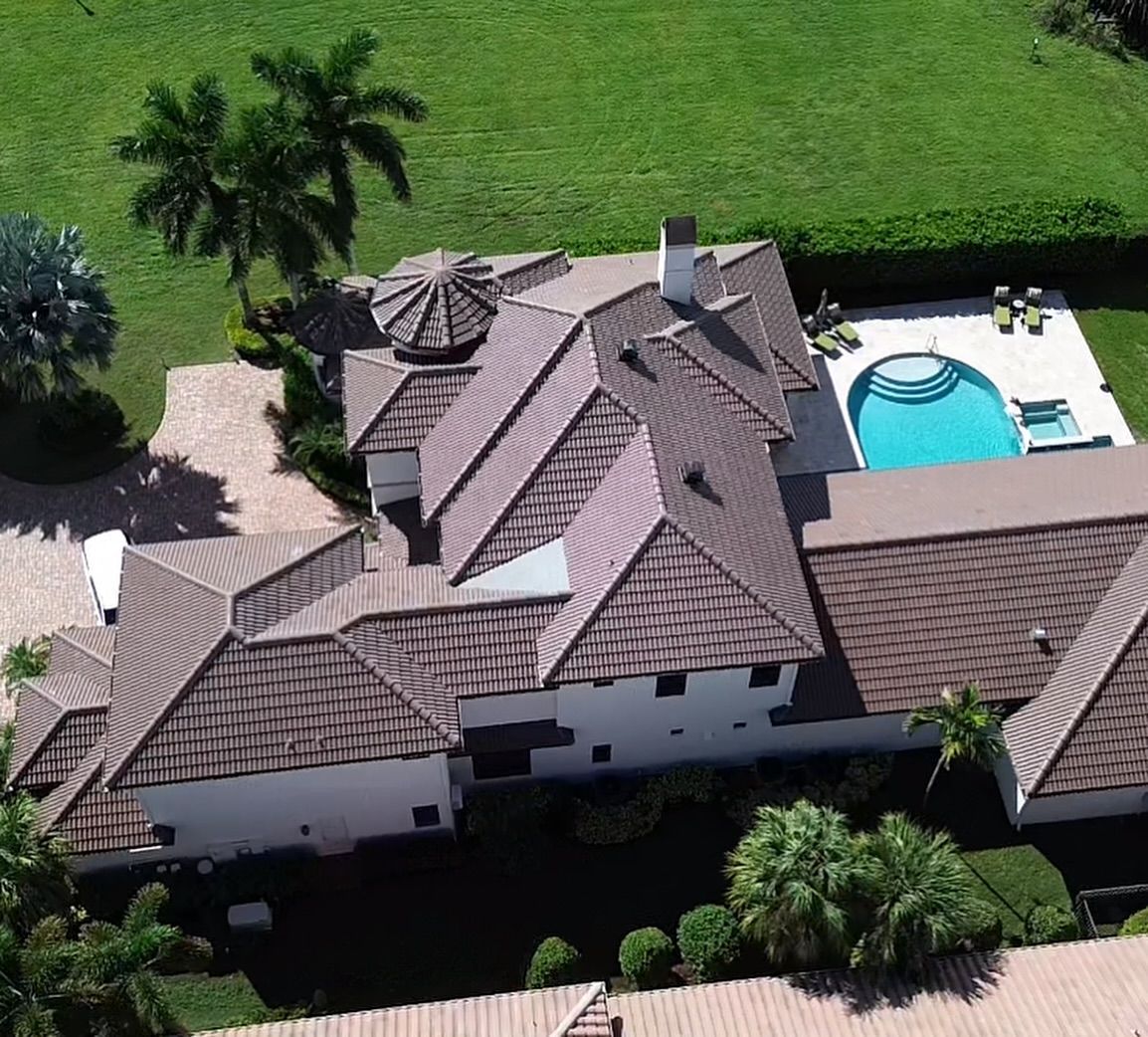 Aerial view of a large white house with a multi-gabled brown roof, a pool, and surrounding green lawn and palm trees.