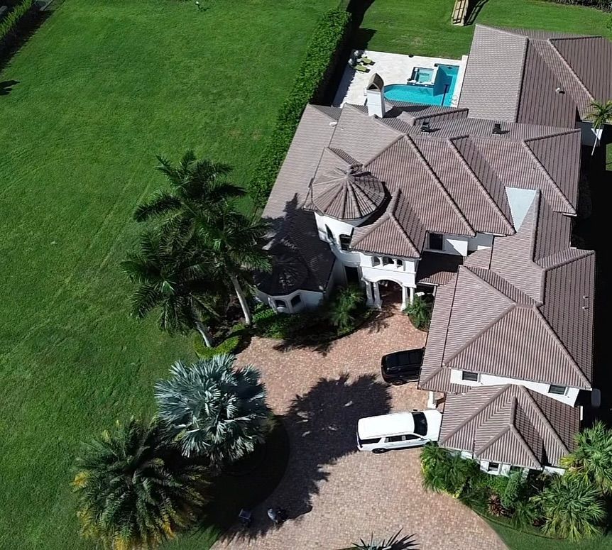 Aerial view of a large white house with a brown tile roof, a swimming pool, and a large lawn with palm trees.