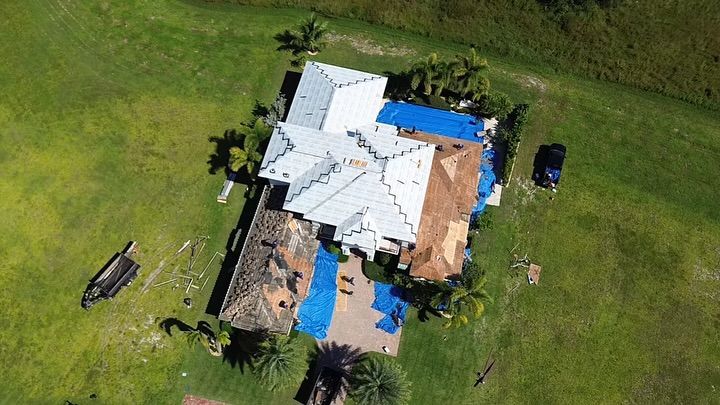 An aerial view of a house under repair, featuring sections of new white roofing, blue tarps, and exposed wooden framing.