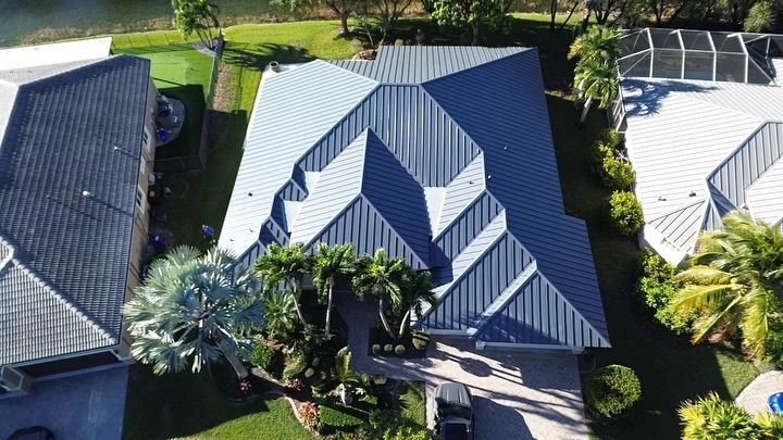 An aerial view of a blue metal roof on a suburban house surrounded by green trees and neighboring homes.