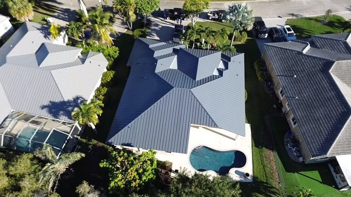 An aerial view shows a house with a dark gray metal roof, a backyard pool, and surrounding neighboring homes and trees.