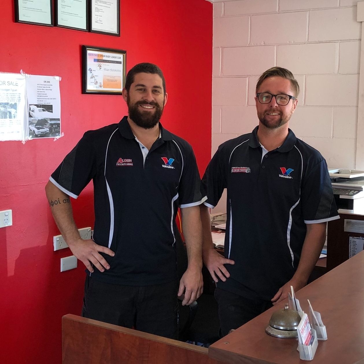 Two smiling staff members in black shirts stand behind a counter in a red-walled office.