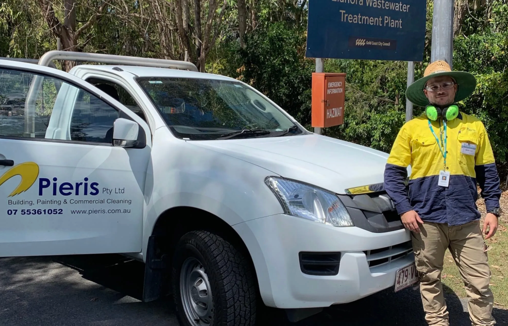 White Pieris utility truck parked by a sign, with a worker in a yellow jacket standing beside it outdoors.