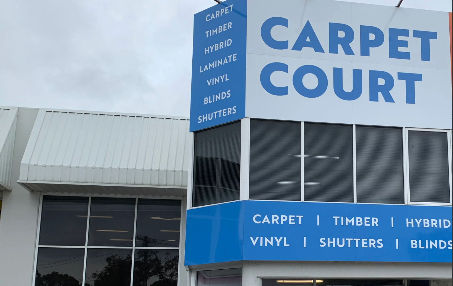 Storefront sign for Carpet Court with blue and white exterior, showing carpet, timber, vinyl, shutters, and blinds.