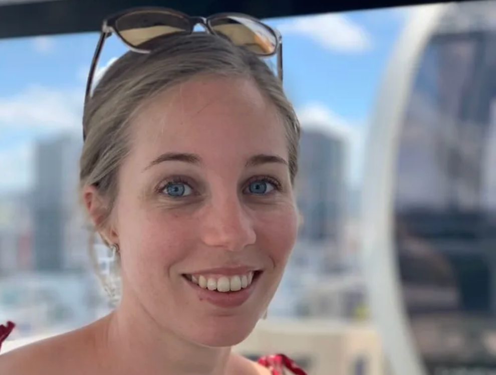 Smiling woman with sunglasses on her head, seated indoors with a blurred city view behind her