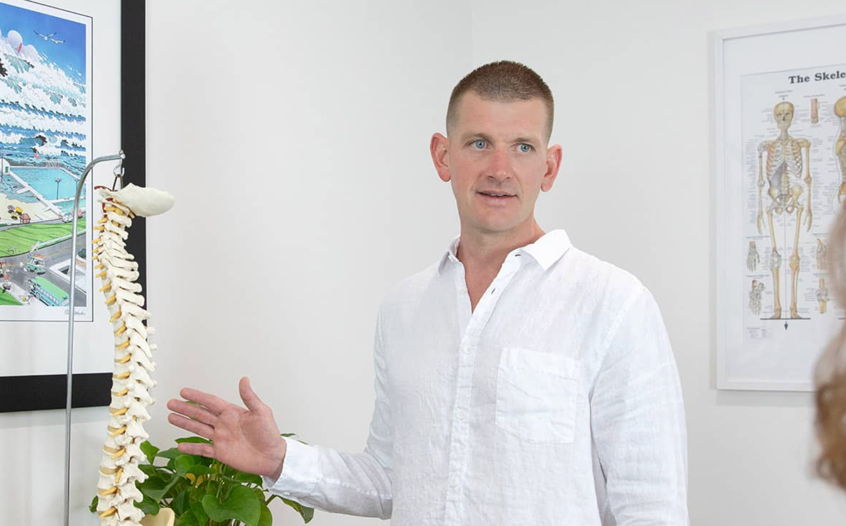 Man in white shirt beside spine model in a clinic, gesturing while standing near anatomical posters.