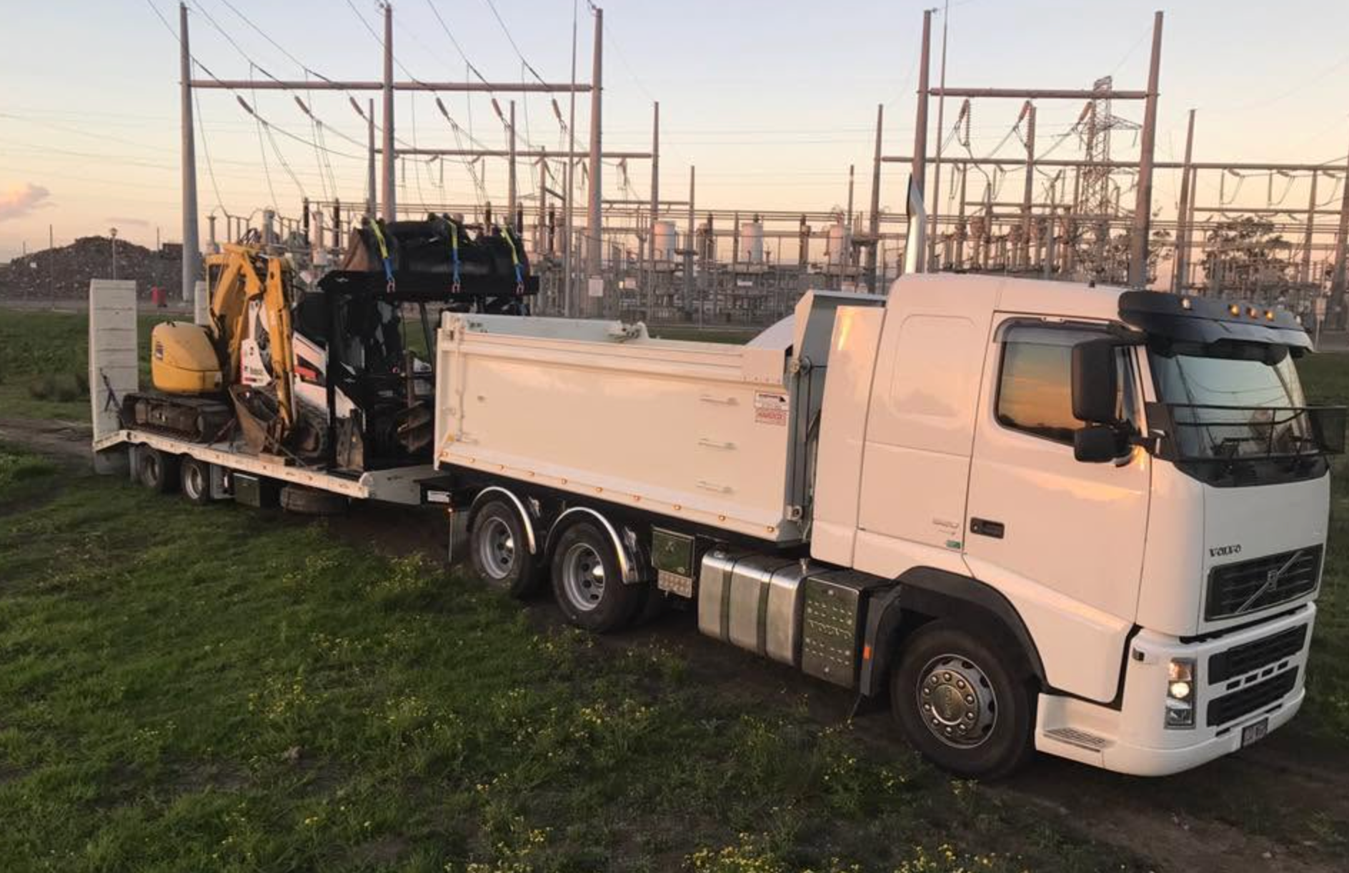 White dump truck and yellow excavator on a low loader at a construction site at sunset