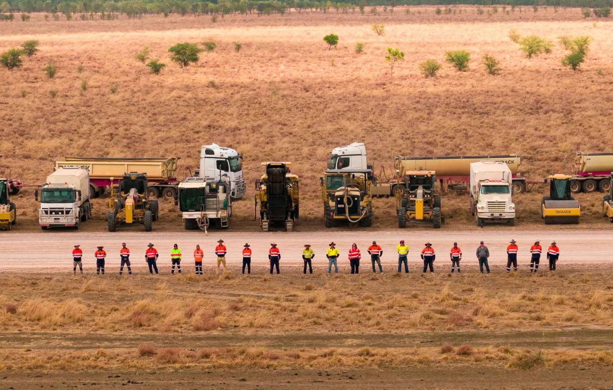 Line of workers in colorful safety vests standing before parked construction trucks on a dry site