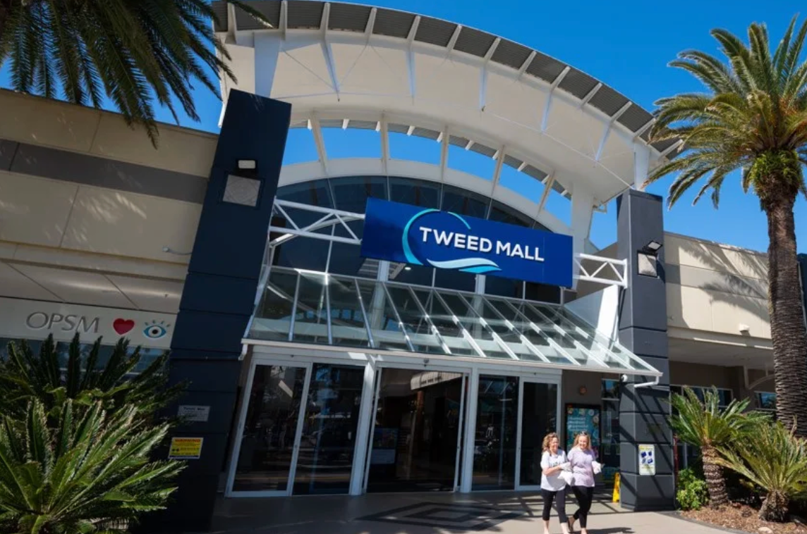 Tweed Mall entrance with blue sign, glass doors, and two people walking outside under a curved awning