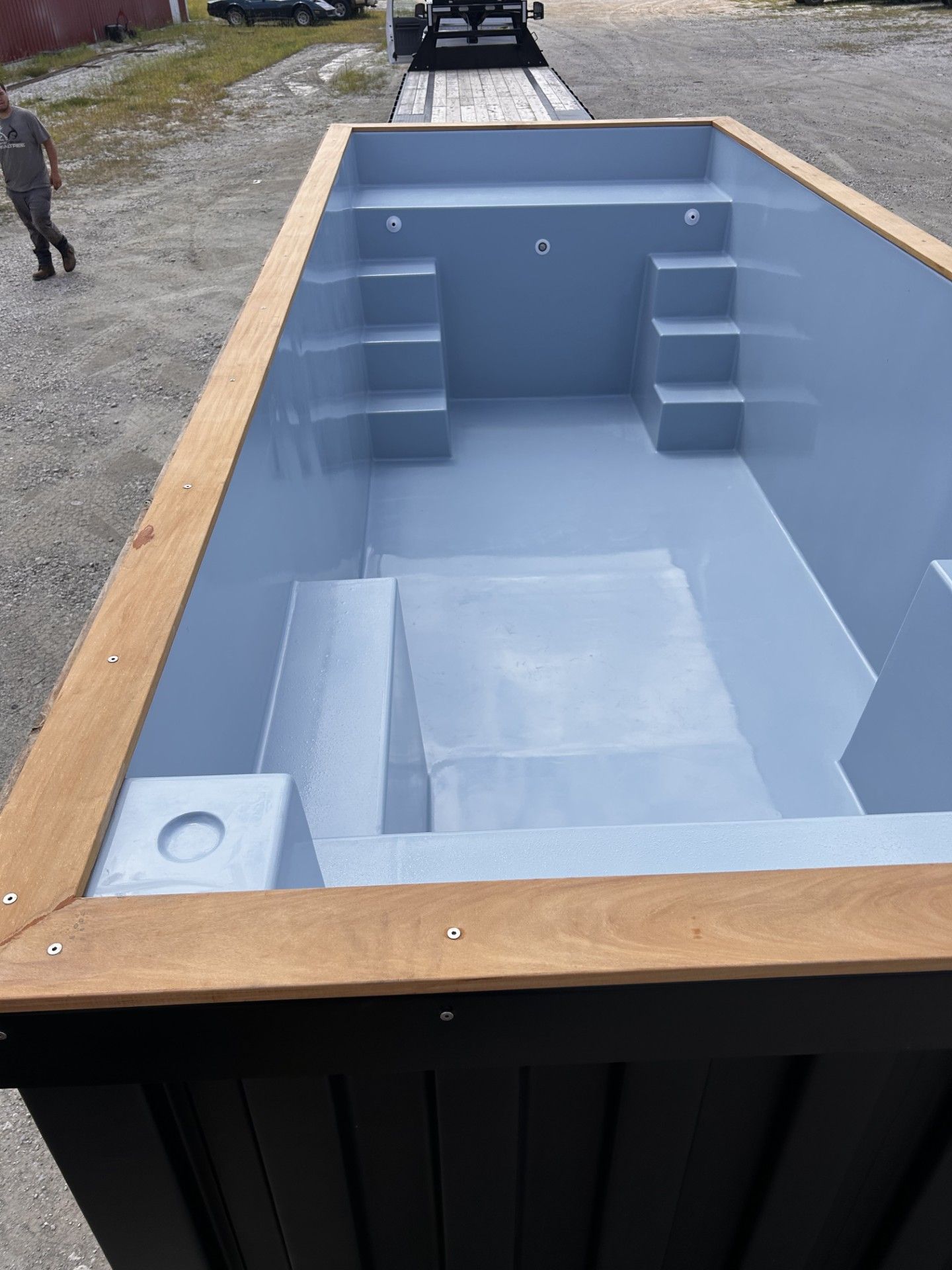 Pre-fabricated light blue swimming pool with wooden border, on a flatbed trailer.