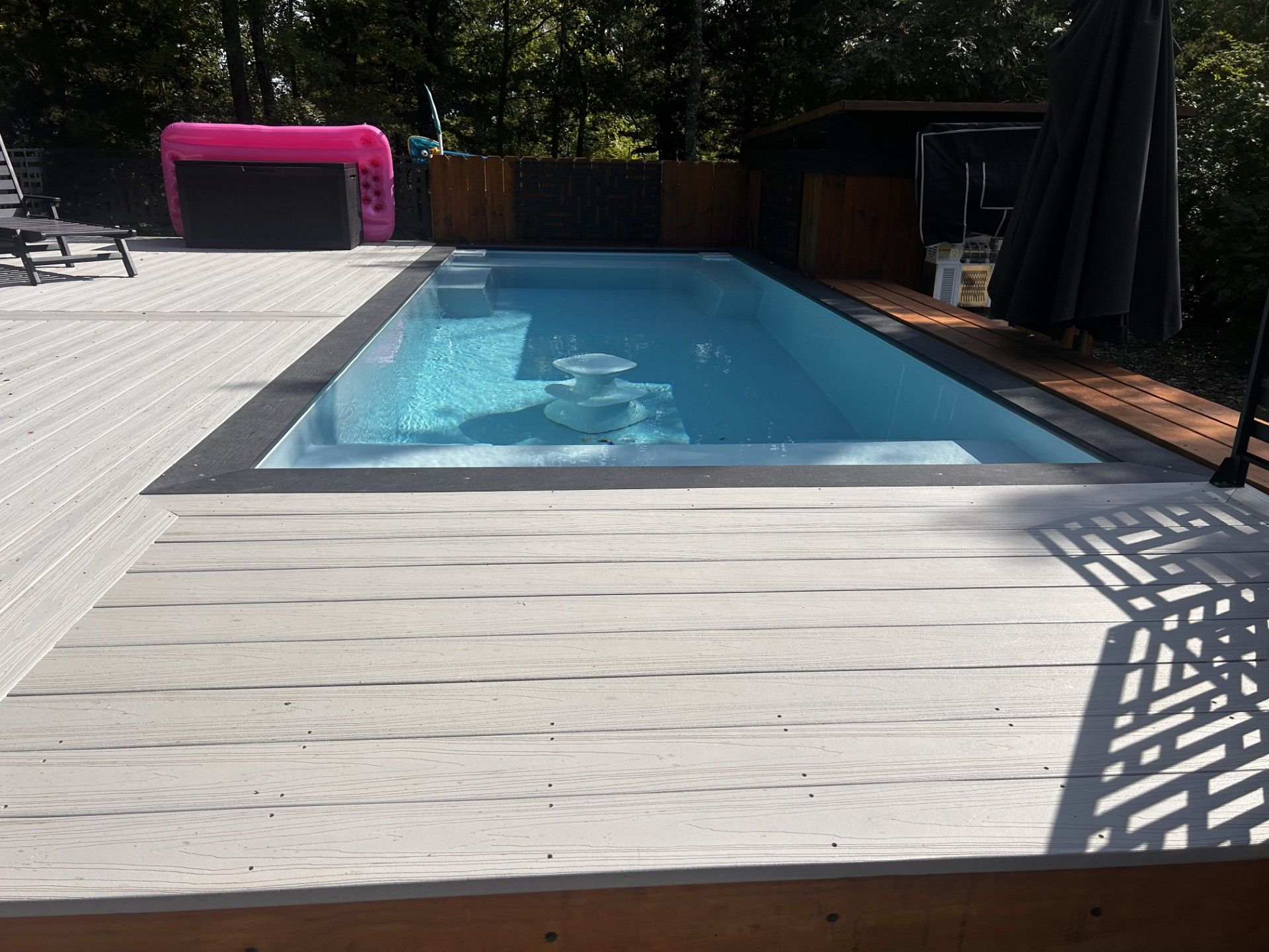Rectangular pool surrounded by gray wooden decking. Pink inflatable toy visible. Trees in the background.