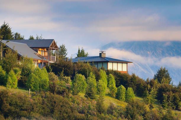 Houses on a hillside with mountain backdrop; trees and green foliage in the foreground under a cloudy sky.