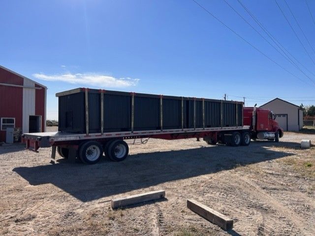 A flatbed semi-truck transporting a large, dark rectangular structure outdoors on a sunny day.