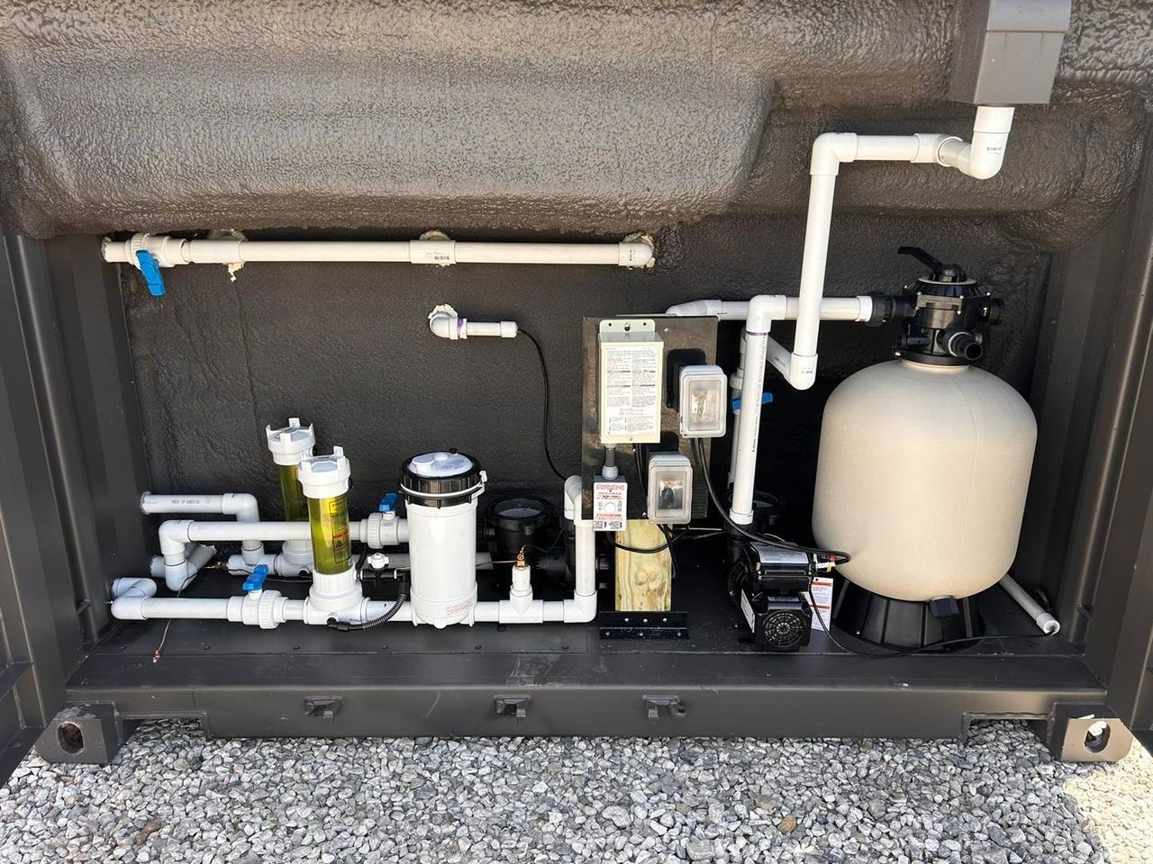 Pool equipment in a dark enclosure: filter, pump, pipes, controls, and a clear view tube.