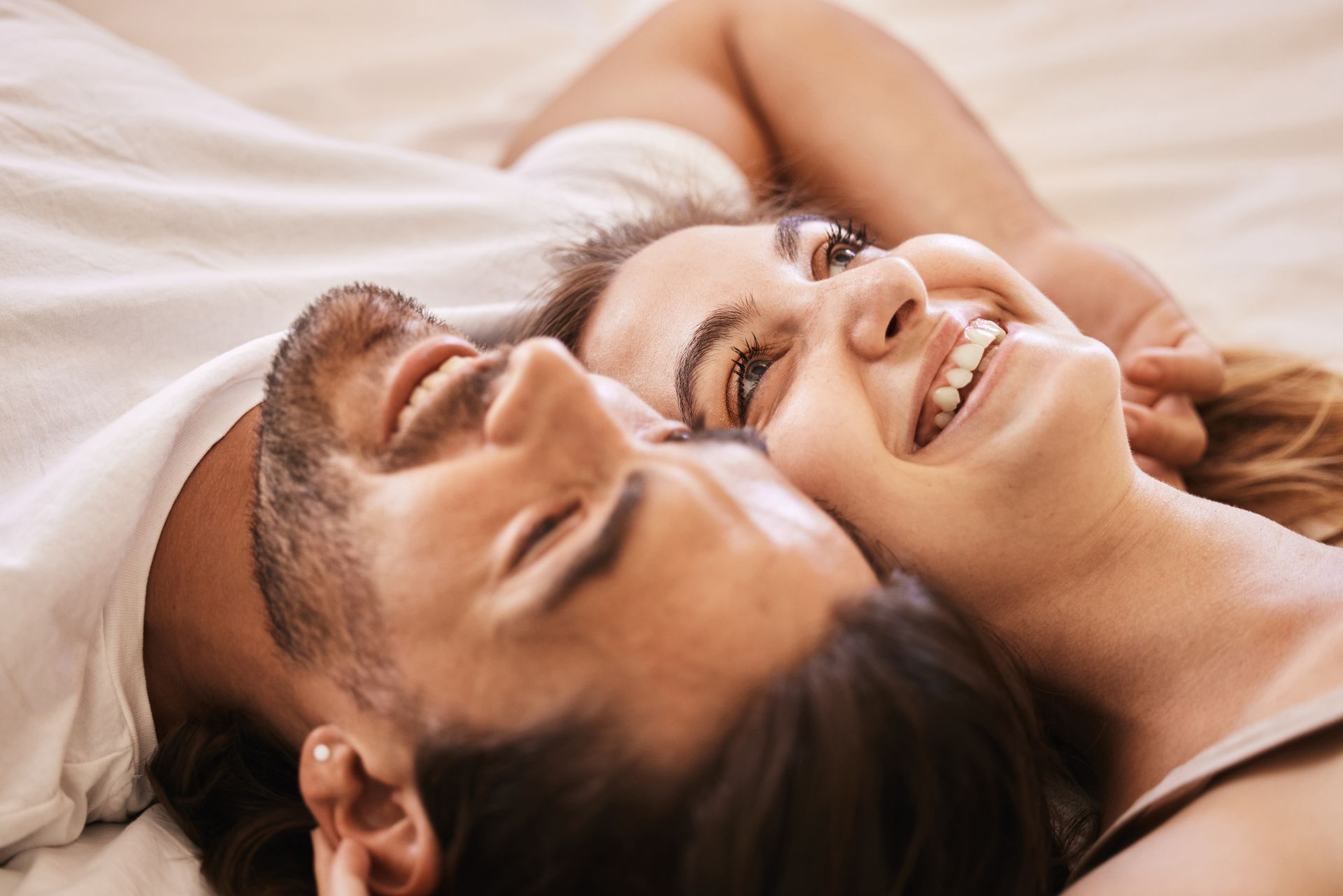 A man and woman lying close together on a bed, smiling warmly while looking upward.