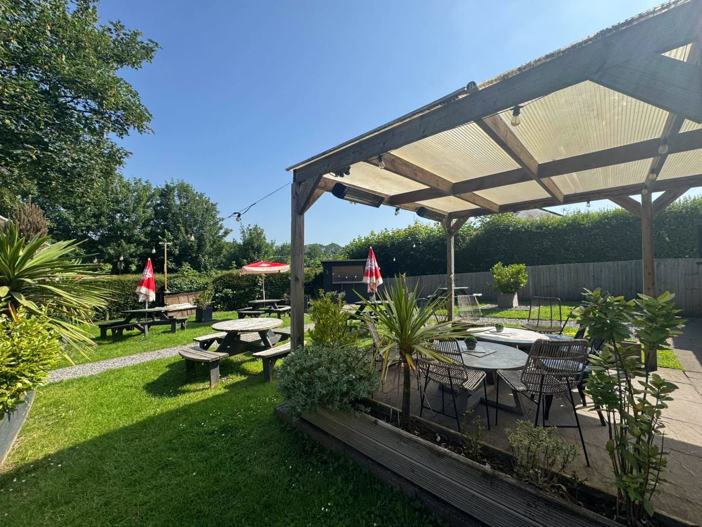 The Bush Inn, St Hilary | Outdoor patio with tables, chairs, and umbrellas under a pergola, surrounded by green lawn & trees.