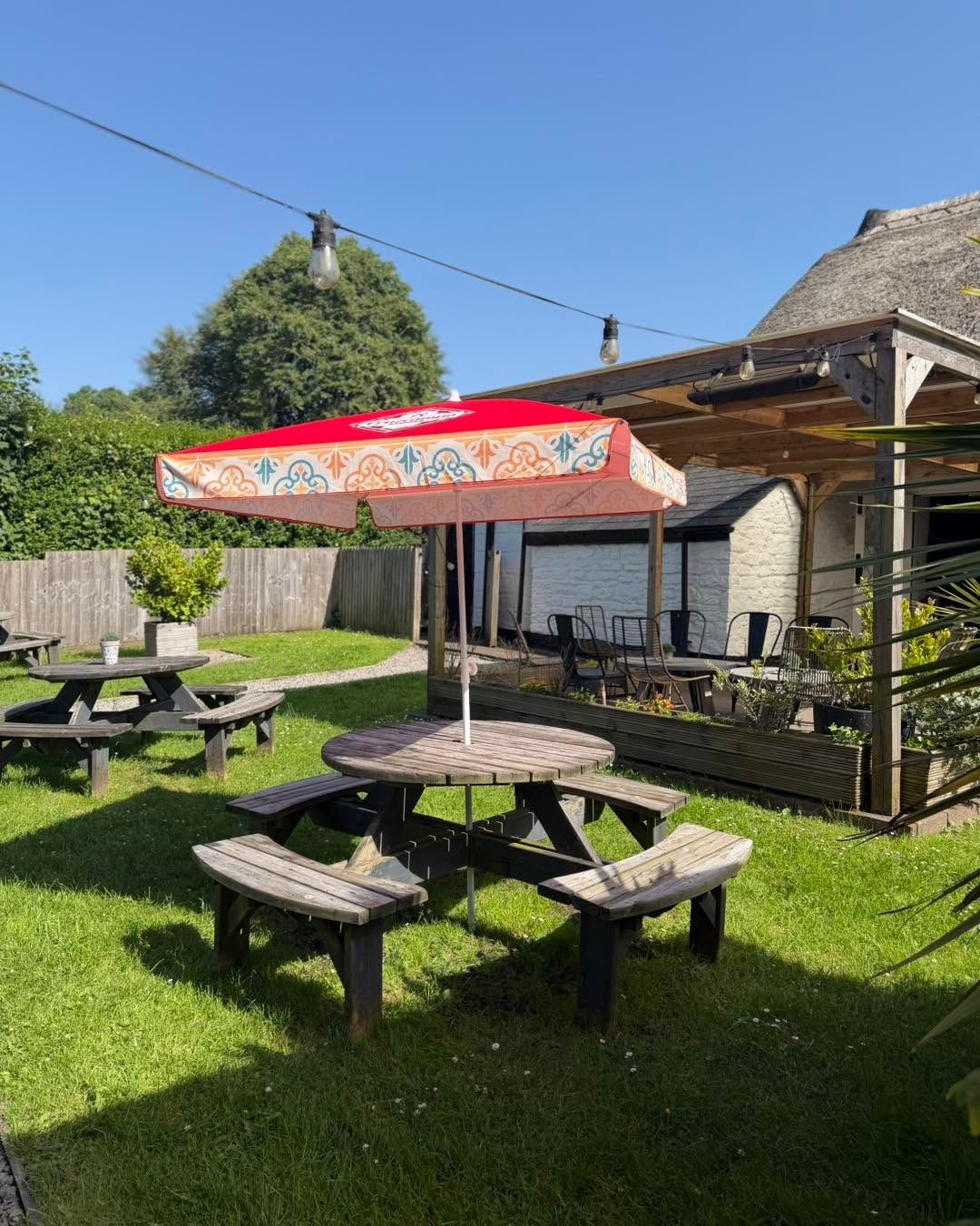 The Bush Inn, St Hilary | Outdoor picnic tables with benches under a red umbrella beside a rustic house on a sunny lawn