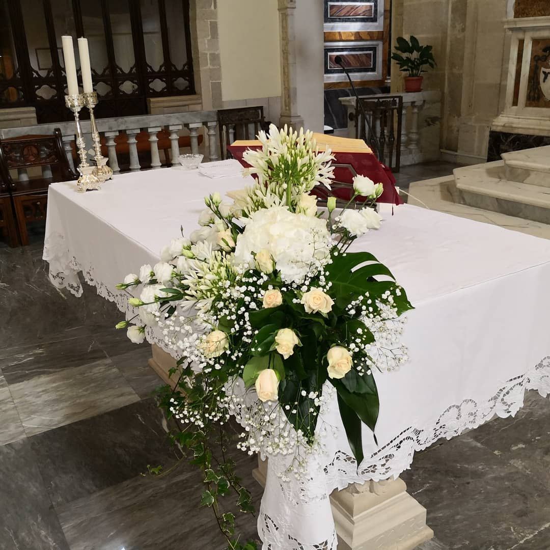 Composizione floreale su un altare bianco in una chiesa, con candele e scalini sullo sfondo.