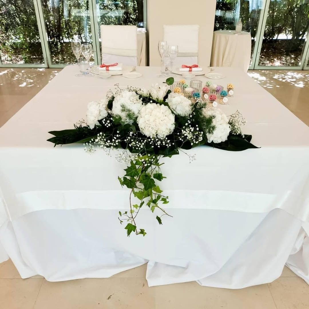 Centrotavola floreale bianco su un tavolo drappeggiato in una luminosa sala eventi al coperto.
