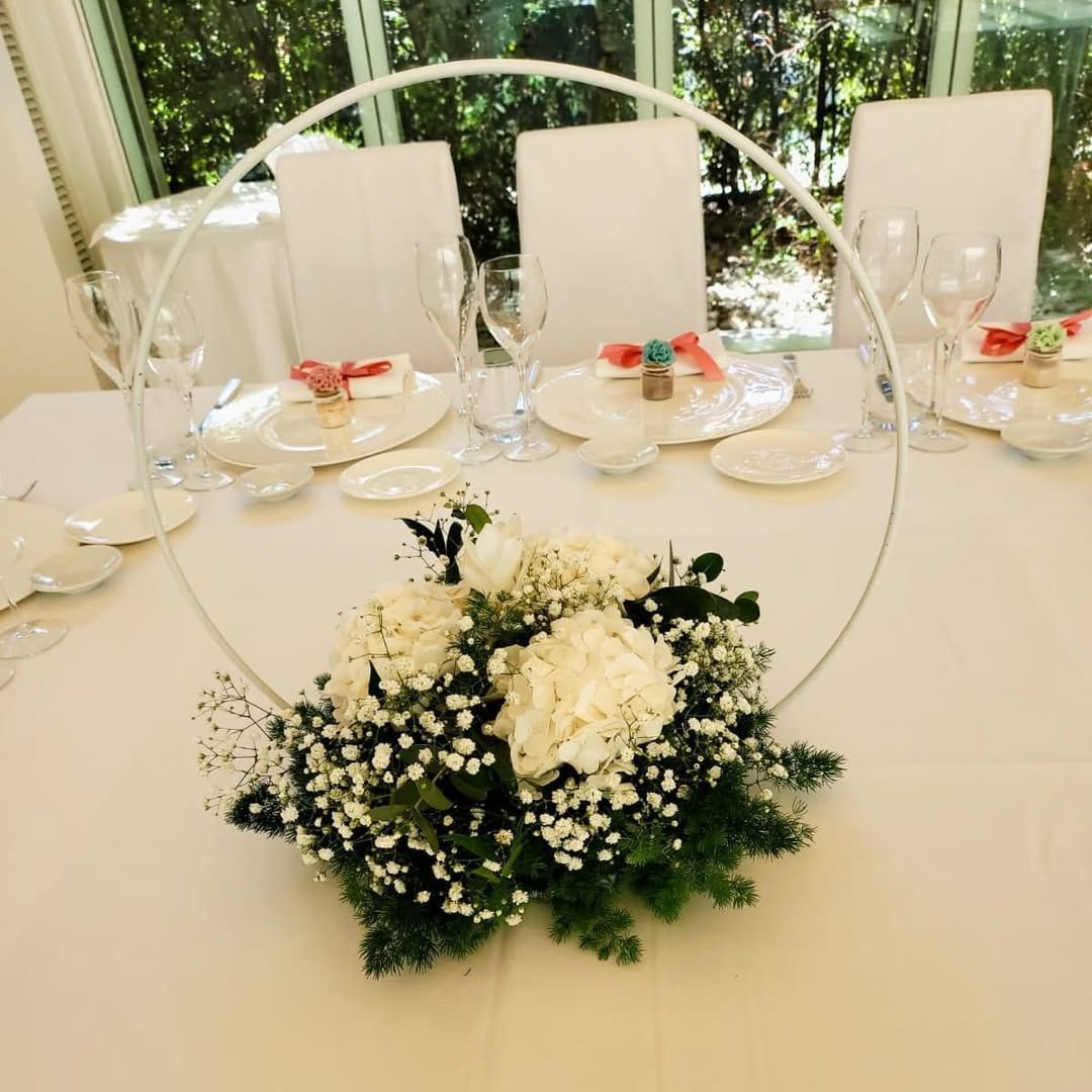 Elegante apparecchiatura della tavola con centrotavola floreale bianco, bicchieri e posate accanto a una finestra luminosa.