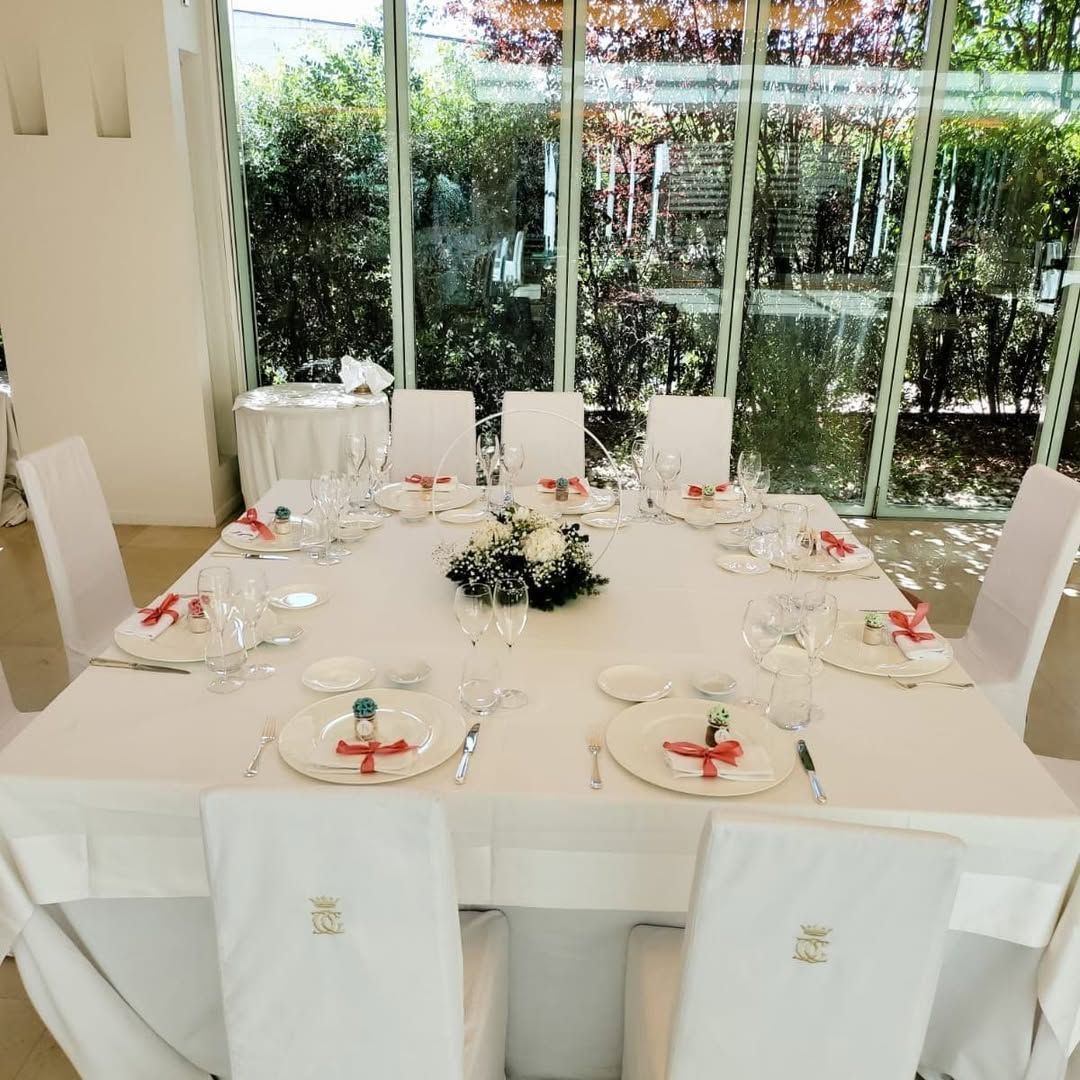 Elegante tavolo da pranzo bianco apparecchiato per un evento formale, con centrotavola floreale e pareti in vetro che si affacciano sul verde.