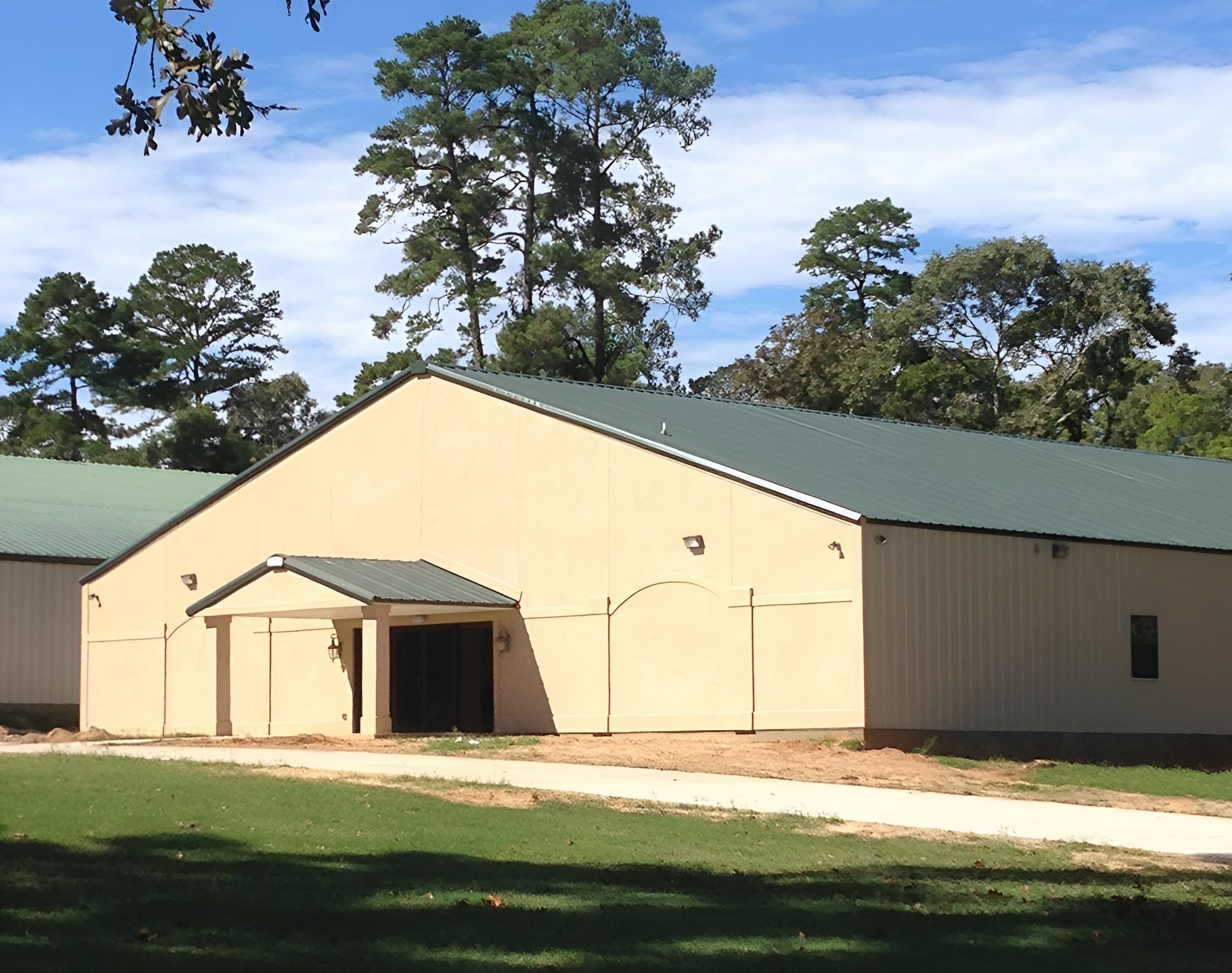 Beige metal building with green roof and small awning, set on grass with trees in the background.
