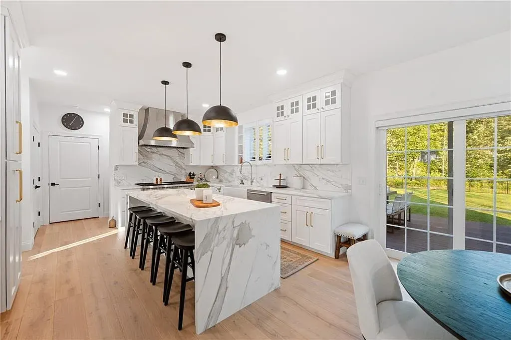 Bright white modern kitchen with marble island, black stools, pendant lights, and sliding glass doors to patio