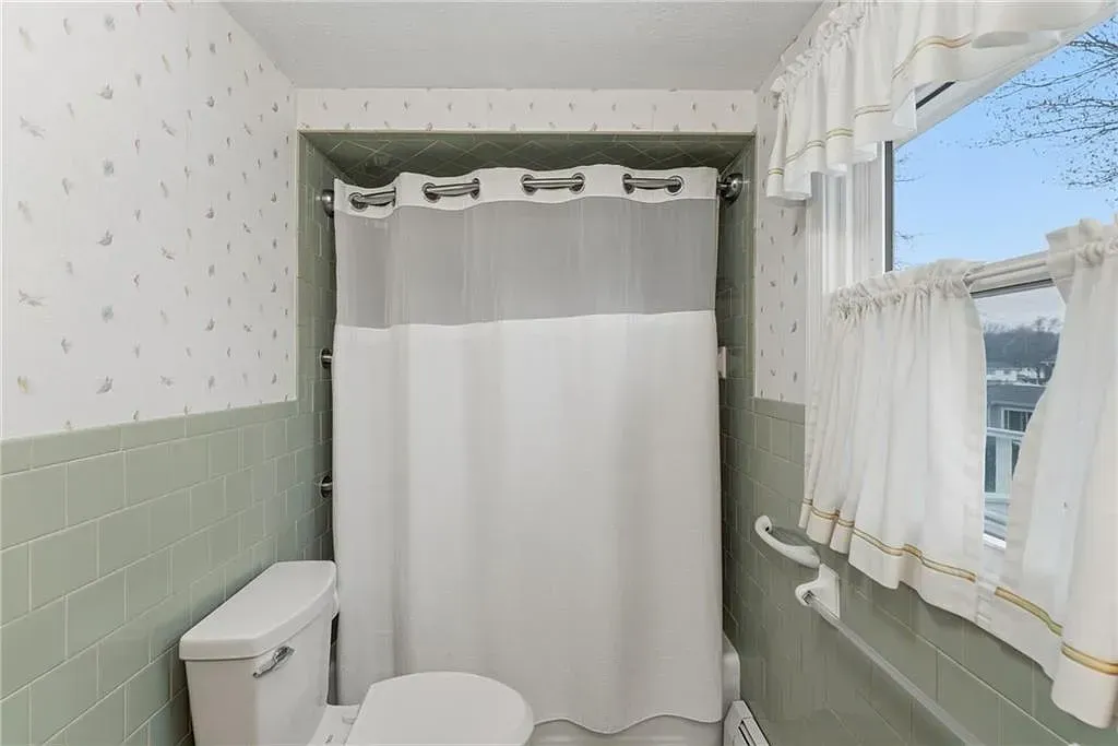 Small bathroom with white shower curtain, toilet, mint-green walls, and a window with white curtains.