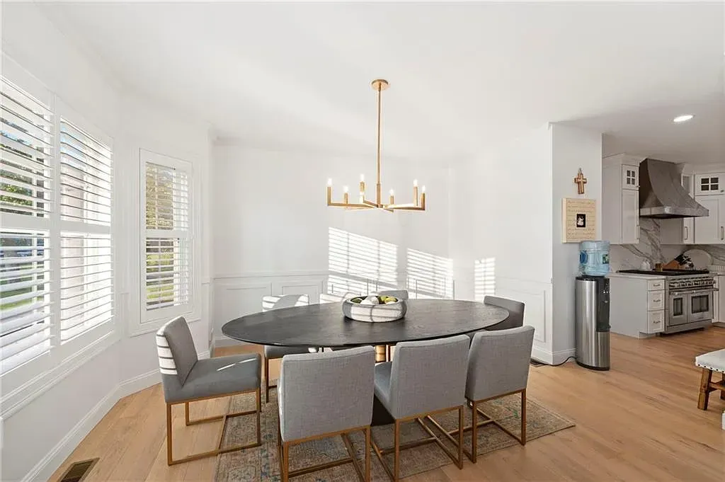 Bright dining room with gray chairs, round table, gold chandelier, and open view into a kitchen
