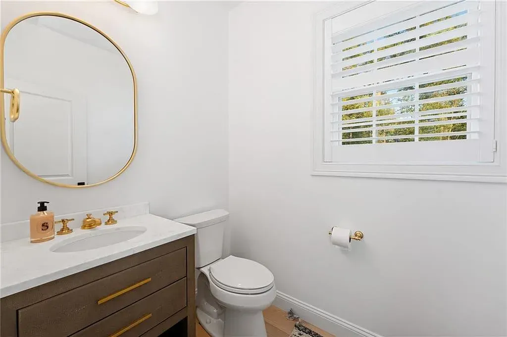 Bright bathroom with a gold-framed mirror, vanity, toilet, and window with white blinds.
