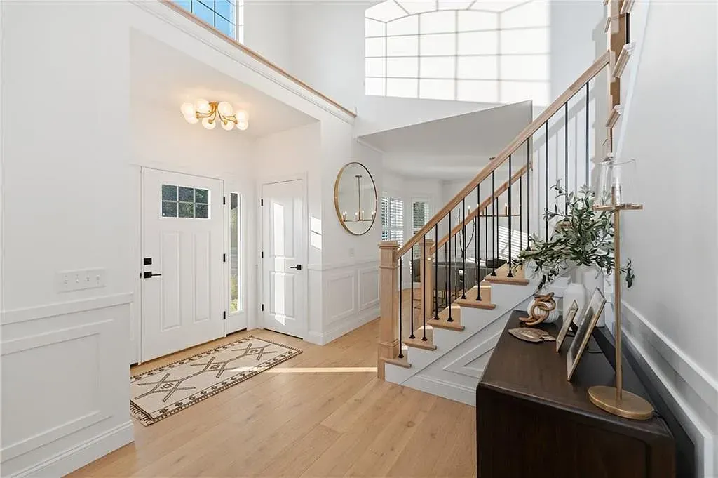 Bright foyer with white walls, wood floors, staircase, front door, and a dark console table with plants.