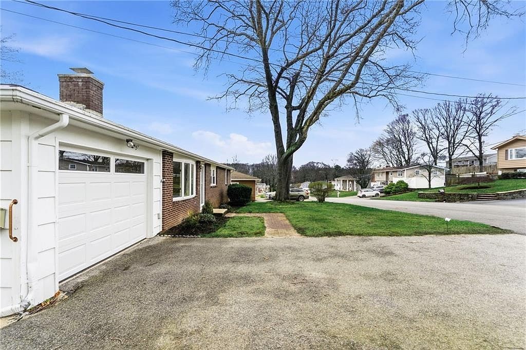 Suburban house with attached garage, gravel driveway, green lawn, and a tree under a blue sky.