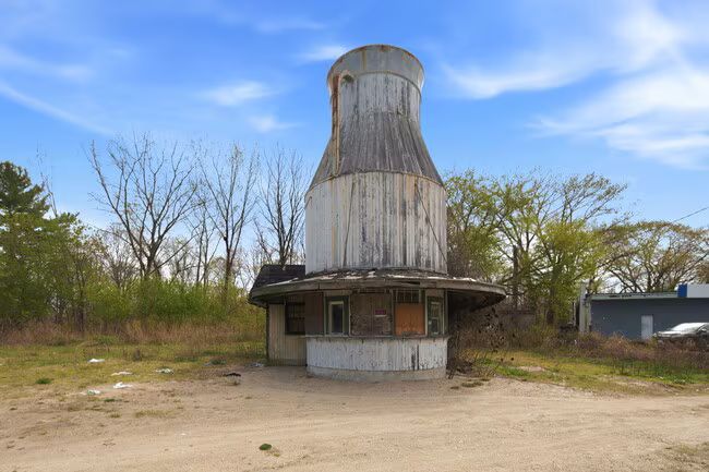 Abandoned concrete tower in a dirt clearing under a blue sky, surrounded by sparse trees.