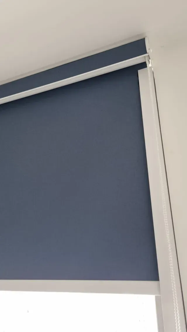 Estor enrollable azul parcialmente bajado sobre una ventana con marco y moldura blancos.