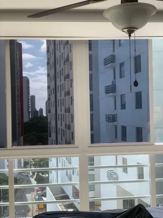 Vista desde una ventana de altos edificios de apartamentos, árboles y una calle abajo, con una lámpara de techo en la parte superior derecha.