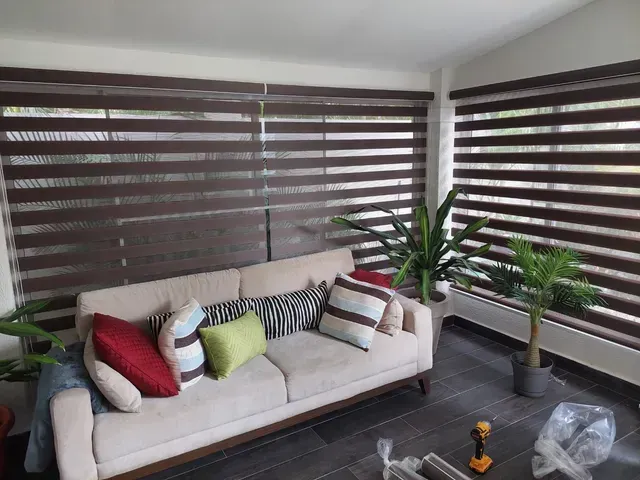 Sala de estar moderna con sofá beige, cojines coloridos, plantas de interior y persianas a rayas.
