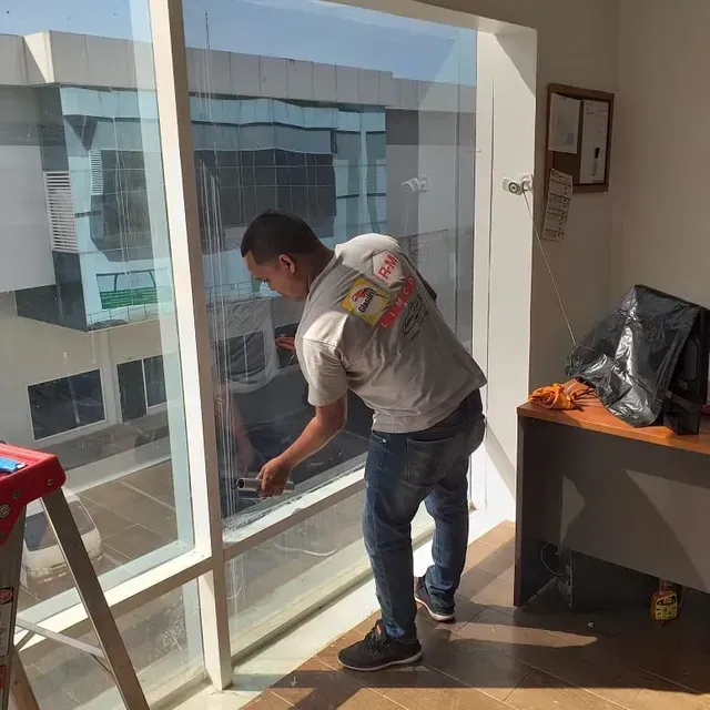 Un hombre limpia una ventana dentro de una oficina, junto a un escritorio y grandes paneles de vidrio.