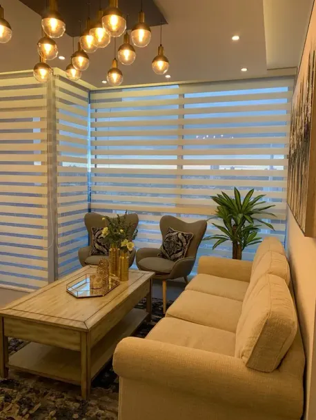 Acogedor salón moderno con sofá beige, mesa de centro de madera, sillones y persianas a rayas bajo lámparas colgantes.