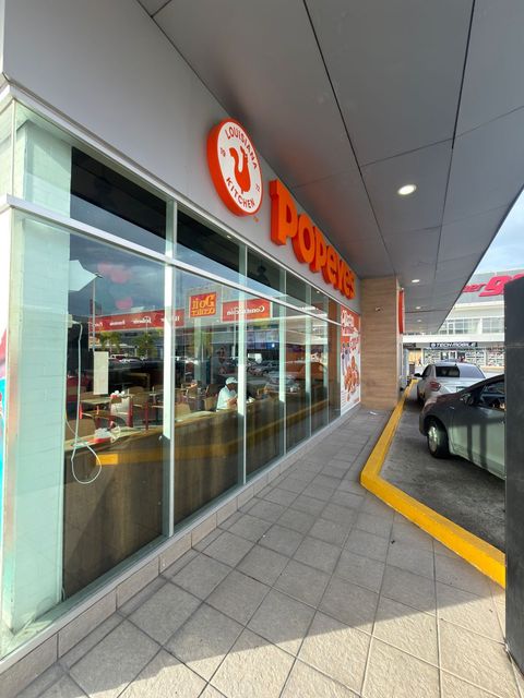 Fachada de tienda con letrero naranja y ventanas de cristal bajo una acera cubierta, con coches aparcados cerca.